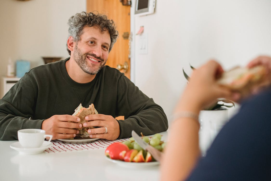 Free Smiling middle aged guy with beard and gray hair in casual wear eating toast while having breakfast with wife in kitchen Stock Photo
